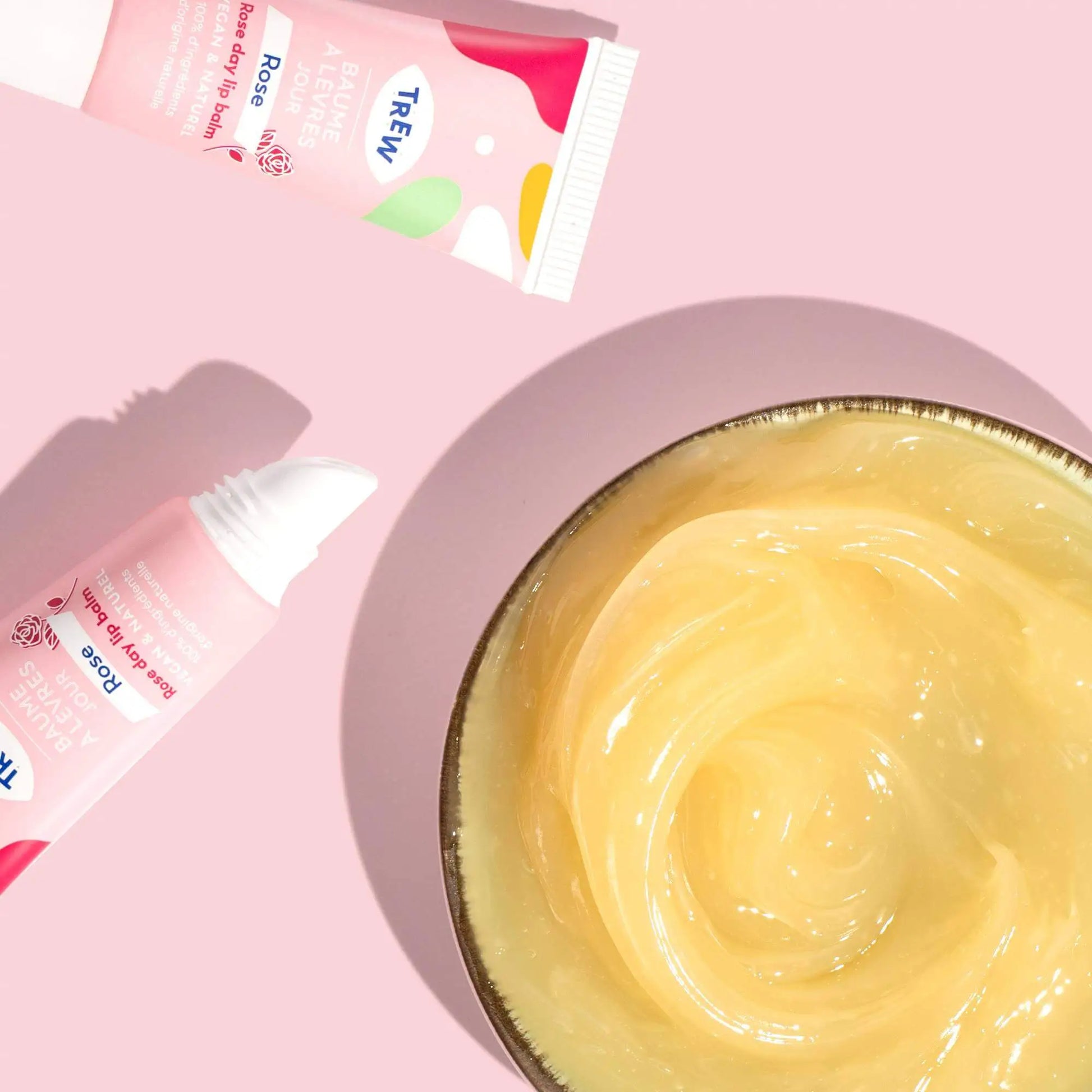 Two tubes of pink Rose lip balm and a small dish filled with golden, smooth balm on a light pink background.