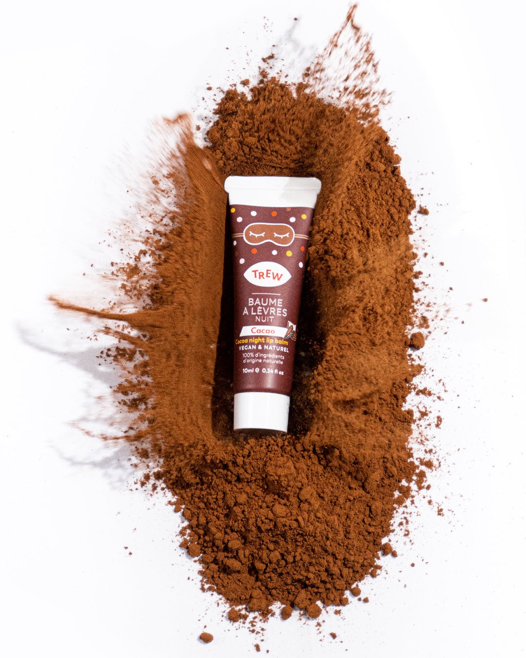 A tube of cocoa night lip balm centered on a splash of brown cocoa powder against a white background.