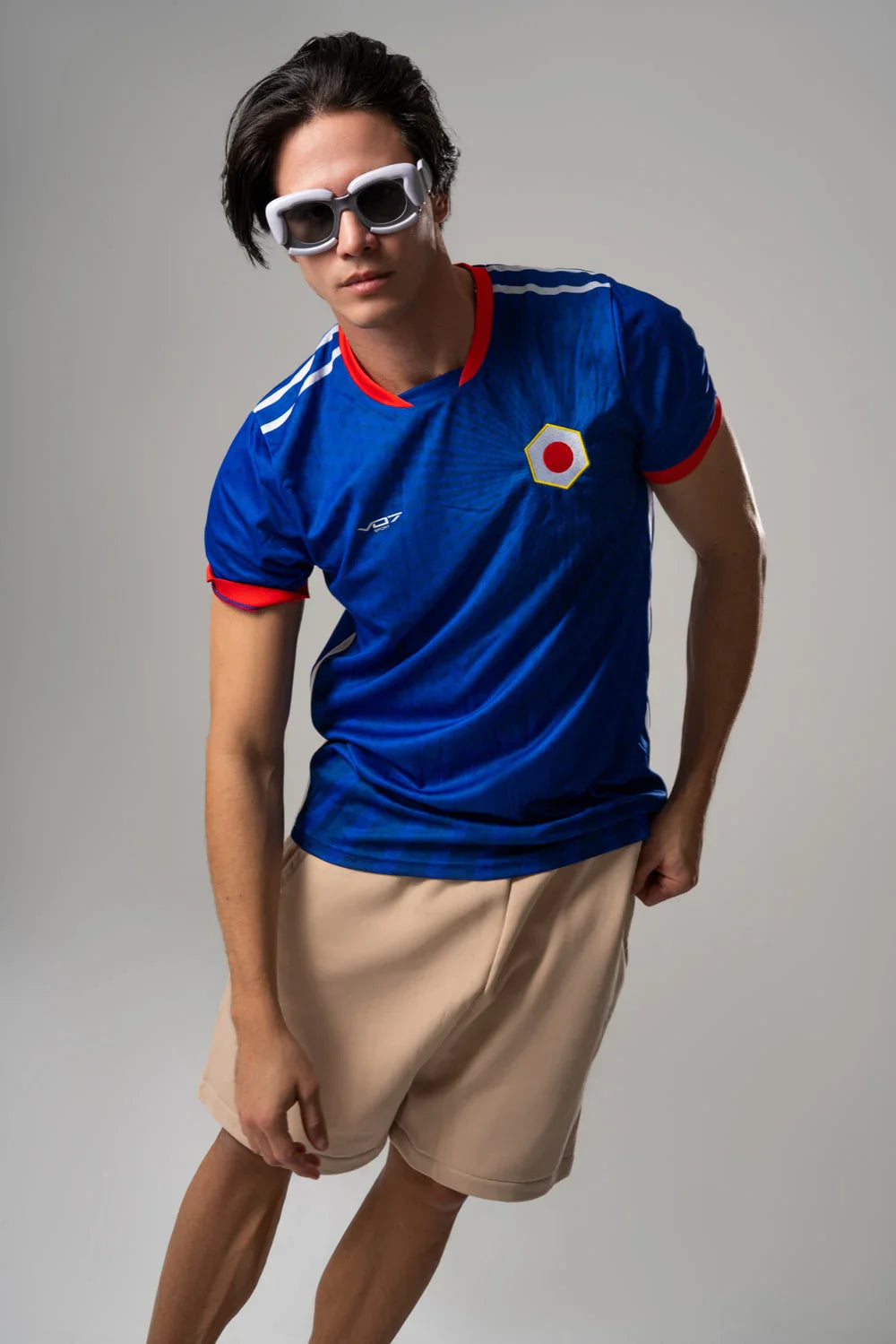 Mannequin portant un maillot VO7 bleu à motifs rayonnants, col et bordures rouges, logo hexagonal et bandes blanches.