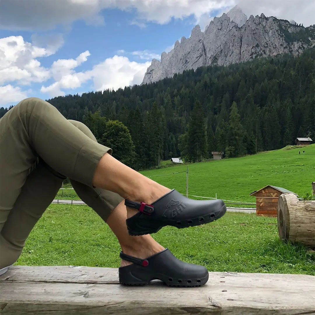 Black slip-on clogs with a heel strap and side cutouts, worn against a scenic mountain landscape.